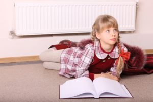 A blonde girl in a red dress with plaid accents and a book in front of her is sitting in front of a white cooling system