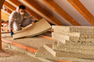 A construction worker wearing a grey jumpsuit and mask is holding a piece of insulation