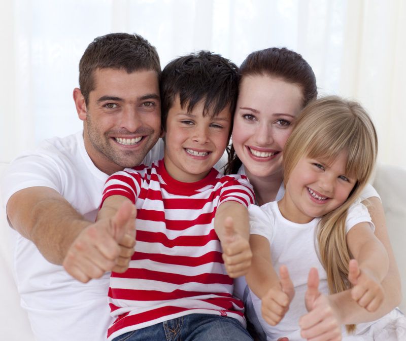 A mother, father, son, and daughter are all smiling and looking at the camera giving a thumbs up. The son is wearing a red and white stripped shirt and the others are wearing white. 