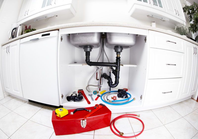 Kitchen sink plumbing with various hoses and tools beneath it. There's a red tool box on the floor. 