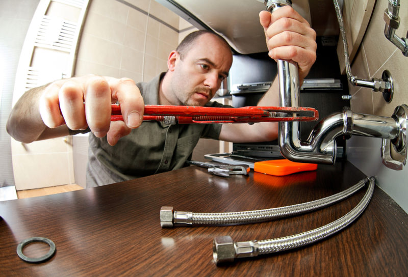 A plumber in a gray shirt using a red wrench to adjust a pipe