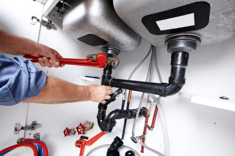 A plumber using a red wrench on a pipe, other wrenches and hoses are on the bottom of the cabinet