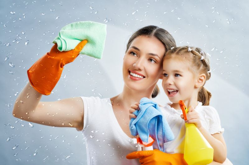 A brunette woman and her blonde child are cleaning a window with a chemical spray cleaner