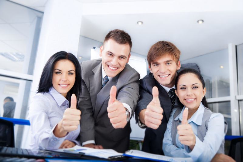 Four office workers are smiling at the camera nd giving a thumbs up. The two women are on the right and left of the two men in the center of the photo. 