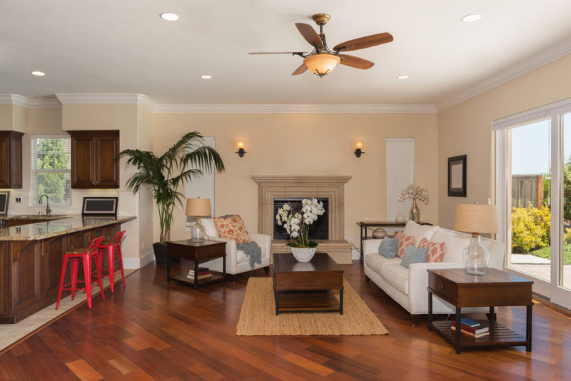 A furnished living room and kitchen countertop with a ceiling fan. There are wood floors, a white couch and chair, and red barstools