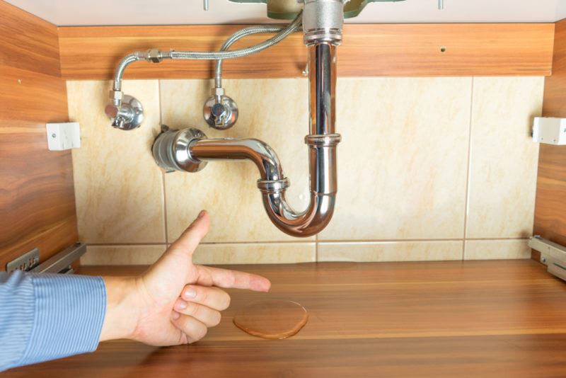 A person pointing to a puddle of water beneath pipes in a wood cabinet